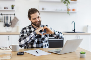 Positive man with loving feelings using internet app showing heart shape to his loving partner....