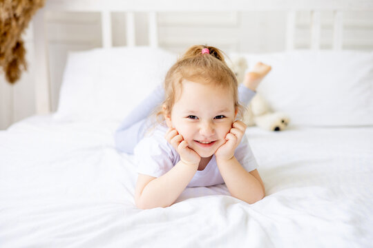 a little cute baby girl on a white cotton bed at home is indulging and having fun smiling on the bed at home with her hands folded under her cheeks