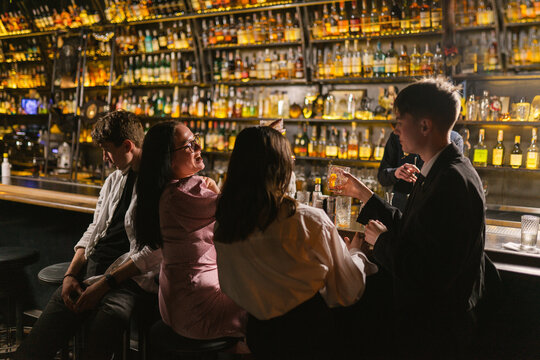 Happy people with cocktails stop noticing friend sitting near at bar counter. Guy feels lonely among young people in pub with dim light