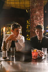 Guy in white shirt sits near sad friend at bar counter in cozy pub. Close friends spend time together at night party in pub