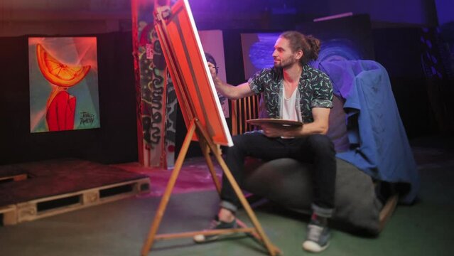 Full length portrait of ingenious male painter sitting in front of wooden easel with canva on it and drawing something indoors. Successful man wearing stylish shirt involved creativity in art studio.