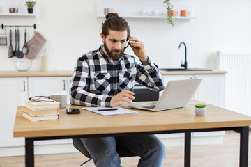 Cheerful adult businessman in wireless headset gesture in front of computer while doing home-based job. Efficient male freelance worker expressing approval of project strategy via video call app.
