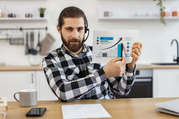 Portrait of Caucasian male in headset posing with infographics on paper at desk with digital devices. Smiling entrepreneur presenting marketing report on online meeting from home workplace.