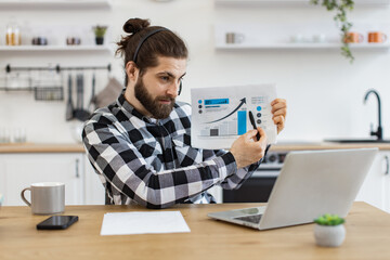 Qualified data analyst demonstrating information about business growth in kitchen interior. Attractive male holding paper with charts while speaking during online meeting via computer at home.