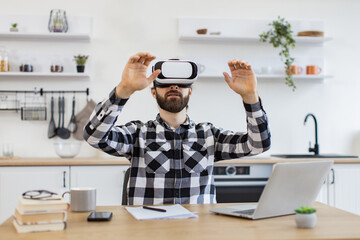 Excited Caucasian worker testing VR headset while sitting at wooden table in modern kitchen. Bearded businessman making presentation using new gadget during remote working online at home.