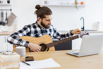 Young male musician in checkered shirt tuning acoustic guitar using laptop on desk in modern...