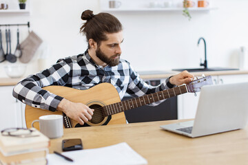 Focused Caucasian male practicing audio exercises on string instrument via online tutorial. Young male musician in checkered shirt tuning acoustic guitar using laptop on desk in modern apartment.