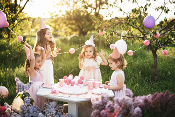 Summer outdoor kids birthday party. Group of happy Children celebrating birthday in park. Children blow candles on birthday cake. Kids party lilac pink pastel decoration and food. Presents and sweets.