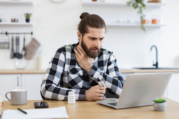 Sick young man in casual clothes touching sore throat while leveraging modern laptop in dining...