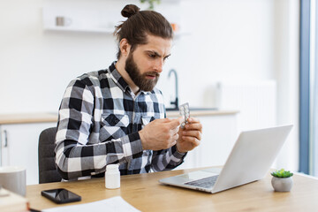 Fototapeta premium Anxious adult person holding round pills while having online conversation via computer on kitchen background. Aching Caucasian man receiving doctor's consultation via internet connection at home.