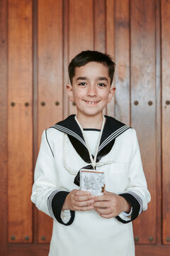A happy boy holding a prayer book, clad in a sailor-style First Communion suit, stands before a wooden door