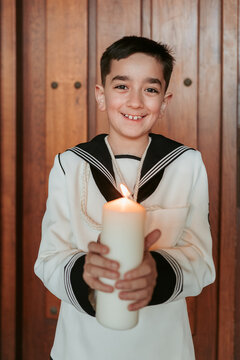 A young boy in his First Communion attire holds a lit candle, symbolizing the light of faith, with a warm smile