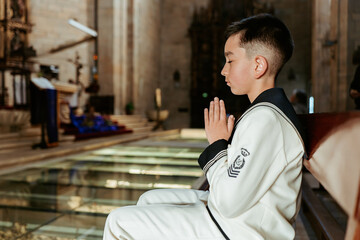 A solemn young boy sits in a church pew, hands clasped in prayer, during his First Communion ceremony, a sacred rite of passage