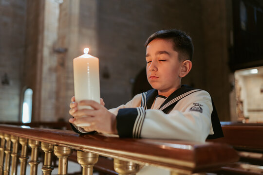 A thoughtful boy holds a lit candle during his First Communion, reflecting in a serene church setting