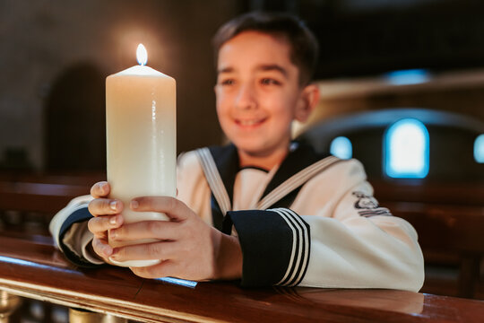 A smiling boy in his First Communion outfit holds a candle with a bright flame, symbolizing joy and faith in the church