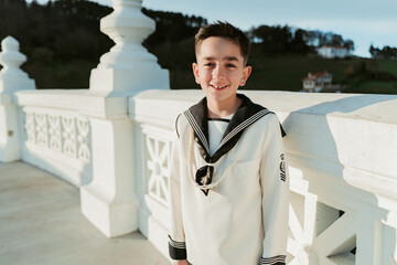 A cheerful young boy wearing his First Communion attire poses with a bright smile on a sunlit terrace, with nature in the backdrop