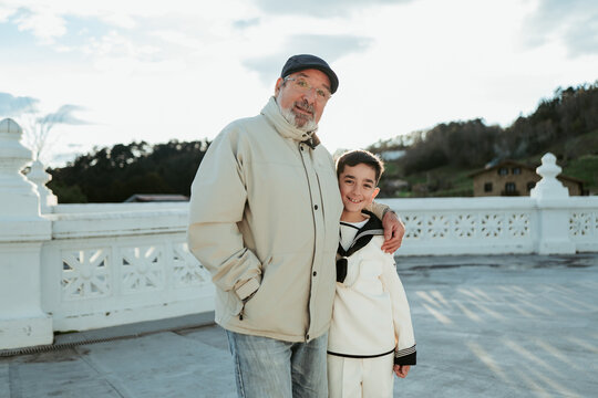 A proud grandfather in a cap embraces his grandson in First Communion attire, sharing a joyful moment on a terrace with a scenic view