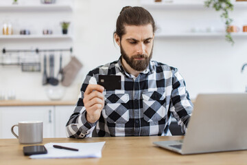 Fototapeta premium Attractive bearded man in checkered shirt holding credit card while typing on portable computer in kitchen interior. Happy Caucasian gentleman doing online banking transaction while working remotely.