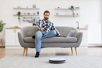 Focus on smart robotic vacuum operated by happy Caucasian man while performing automatic cleaning on carpet indoors. Cheerful owner programming home gadget via remote control while focusing on work.