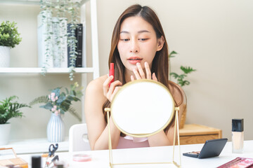 Skin care, beauty cosmetics concept, pretty asian young woman, girl make up face by applying red lipstick, lips balm on her mouth, looking at the mirror at home. Female look with natural fashion style