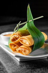 Golden spring rolls tied with a green pandan leaf, served on a white plate with dipping sauce,...