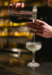 Fototapeta na wymiar Bartender pours white creamy drink through sieve in glamorous glass. Barman makes elite alcoholic beverage by counter in nightclub