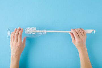 Young adult woman hand holding white brush and washing inside transparent plastic bottle on light blue table background. Pastel color. Point of view shot. Closeup. Top down view.