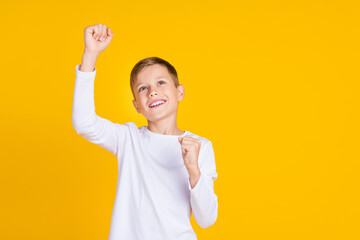 Photo of sweet lucky kid dressed white shirt rising fists looking empty space isolated yellow color...