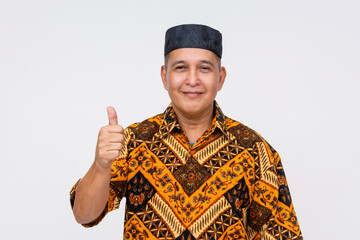 Fototapeta premium Portrait of a cheerful Indonesian man wearing a traditional batik shirt and songkok, giving a thumbs-up gesture on a white background.