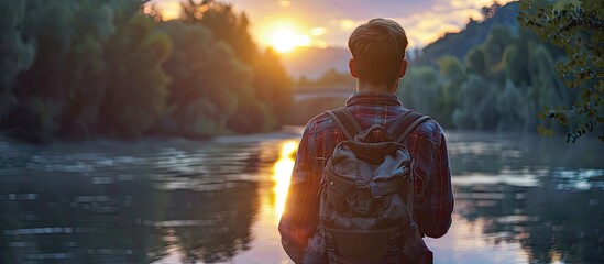 A happy traveler with a backpack stands by the water at sunset, admiring the picturesque landscape with trees and colorful clouds in the sky