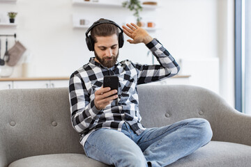 Passionate young man wearing checkered shirt enjoying loud music in headphones while relaxing in dining room. Happy Caucasian adult using cell phone application for tracks download indoors.