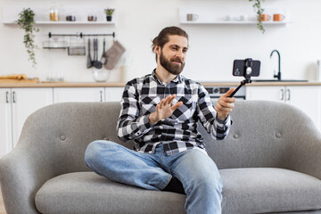 Attractive young man holding phone and selfie stick while talking via webcam on kitchen. Happy...