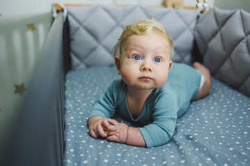 A 6-month-old child lies in a crib and looks at the camera. Gray bed linen for a baby bed