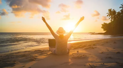 Fototapeten A woman sitting on the beach with a laptop, her arms raised in joy and celebration of success. A beautiful sunset at a tropical island in the background. A concept for a freedom lifestyle  © Olya Fedorova