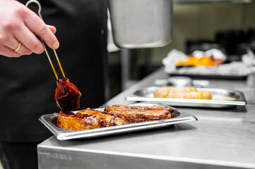 Professional chef in a kitchen glazing freshly grilled ribs with a thick sauce, with other food trays in the background.