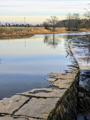 Spring Riverside Reverie: A Gently Flowing River Cascades Over a Weathered Stone Weir, Mirroring a Tranquil Sky and tree at before sunset