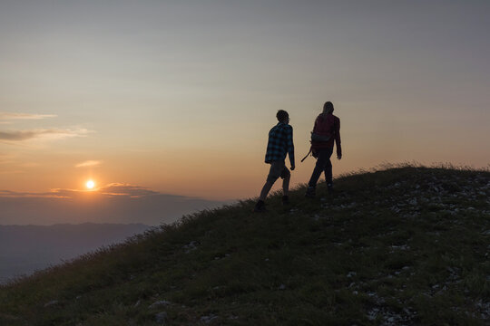 Two silhouettes, male and female, conquering a mountain peak, giving high fives and enjoying the landscape during a romantic sunset, aerial shot.