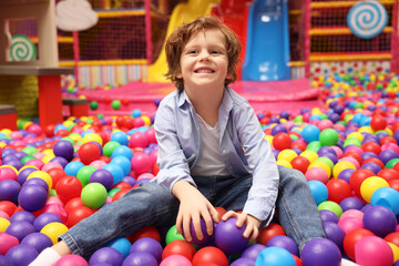 Happy little boy sitting on colorful balls in ball pit