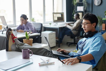 Side view portrait of Asian young man working with laptop while relaxing casually at workplace in...