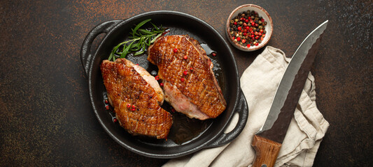Two roasted duck breast fillets with crispy skin, with pepper and rosemary, top view in black cast iron pan with knife, dark brown concrete rustic background. - 761502313