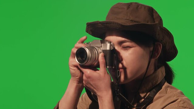 Young female tourist wearing khaki outfit and hat photographing nature with digital camera on green chroma key background