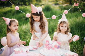 Summer outdoor kids birthday party. Group of happy Children celebrating birthday in park. Children blow candles on birthday cake. Kids party pink pastel decoration and food. Presents and sweets. - 761510177