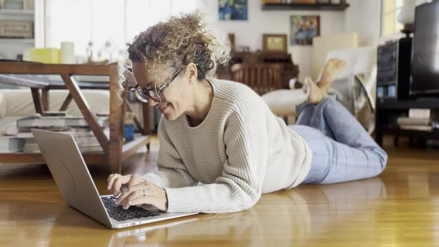 Smiling mature woman laying down on the floor using modern laptop browsing unlimited wireless internet, happy young woman freelancer work on computer typing texting from home, technology concept
