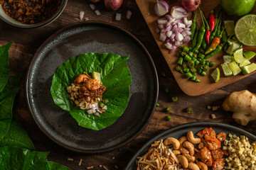 Miang Kham. Thai herbal appetizer wrapping ingredients consist of dried shrimp, roasted coconut, beans, ginger, lime, chili, and herbs into fresh Betel leaves on a wood background.