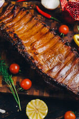 roasted pork ribs in a smokehouse, standing on a cutting board, garnished with rosemary, pepper, chili pepper, tomato, lemon, pomegranate. aromatic baked meat
