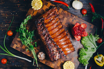 roasted pork ribs in a smokehouse, standing on a cutting board, garnished with rosemary, pepper, chili pepper, tomato, lemon, pomegranate. aromatic baked meat