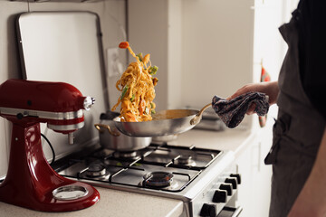 Person flipping pasta in pan on stove, dynamic home cooking scene with kitchen mixer