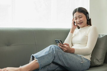 Cropped view of cheerful, carefree Asian woman singing and listening to music on smartphone app using wireless headphones Satisfied smile sitting on the sofa at home On a casual day of the holiday