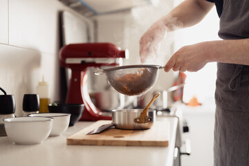 A person cooking in a kitchen, straining shrimps over a pot, steam rising.