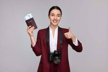 Smiling businesswoman with passport, tickets and camera showing thumb up on grey background